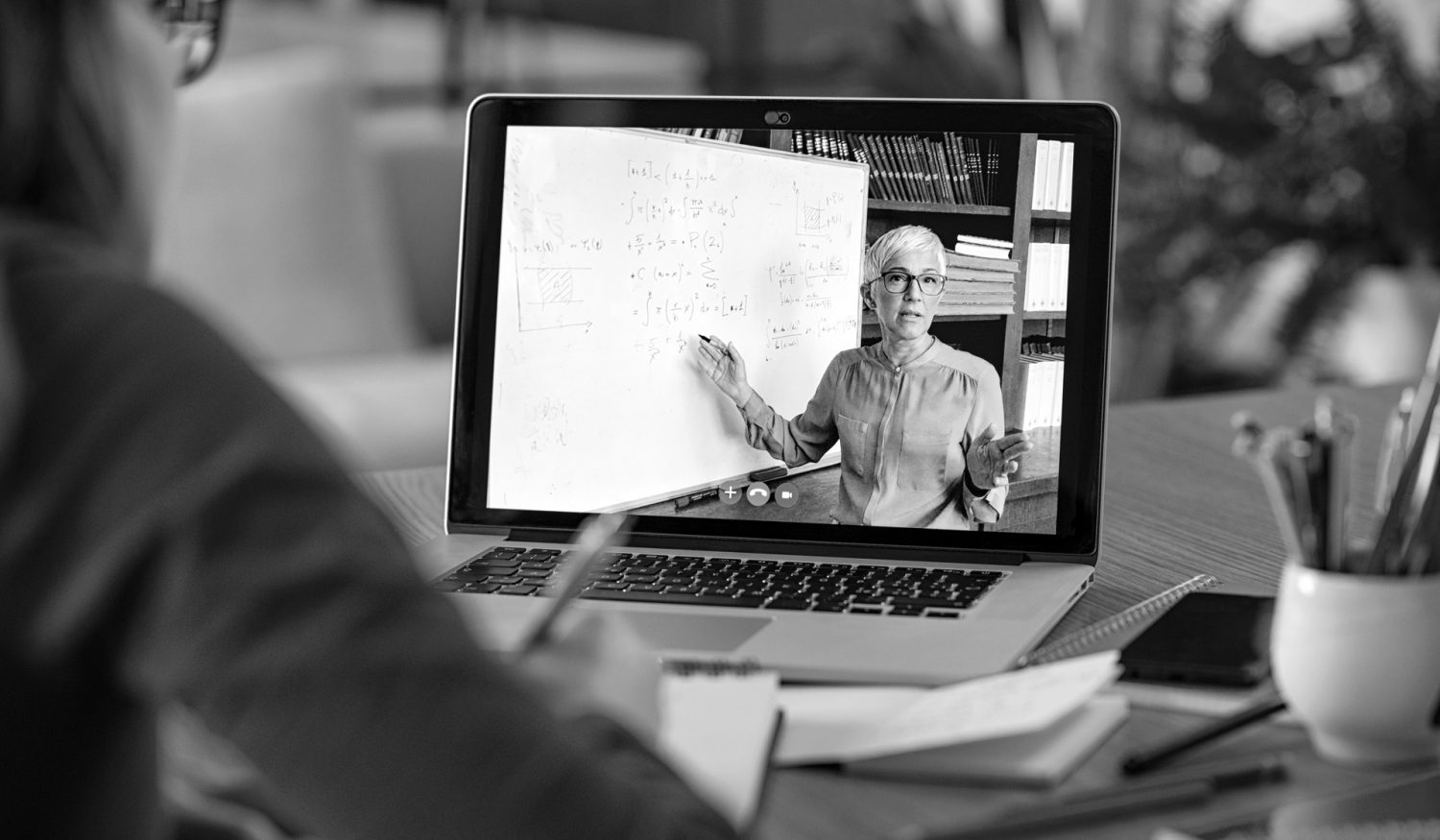 Young student watching lesson online and studying from home. Young woman taking notes while looking at computer screen following professor doing math on video call. Latin girl student studying from home and watching teacher explaining math formula on video chat.