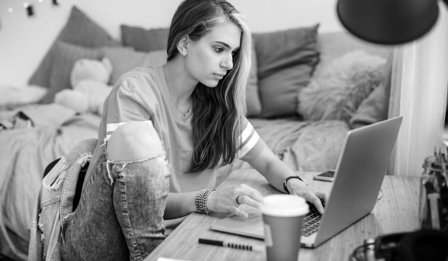 Young woman alone in bedroom. She is working homework, using laptop, sitting on beed and holding teddy bear, drinking coffee, and writing on papper